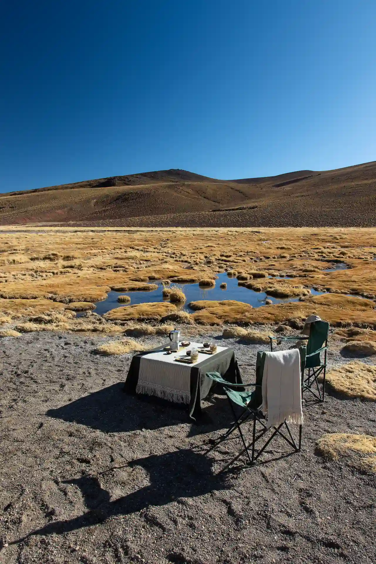 Outdoor table and chairs set beside a high-altitude desert wetland under a clear blue sky.