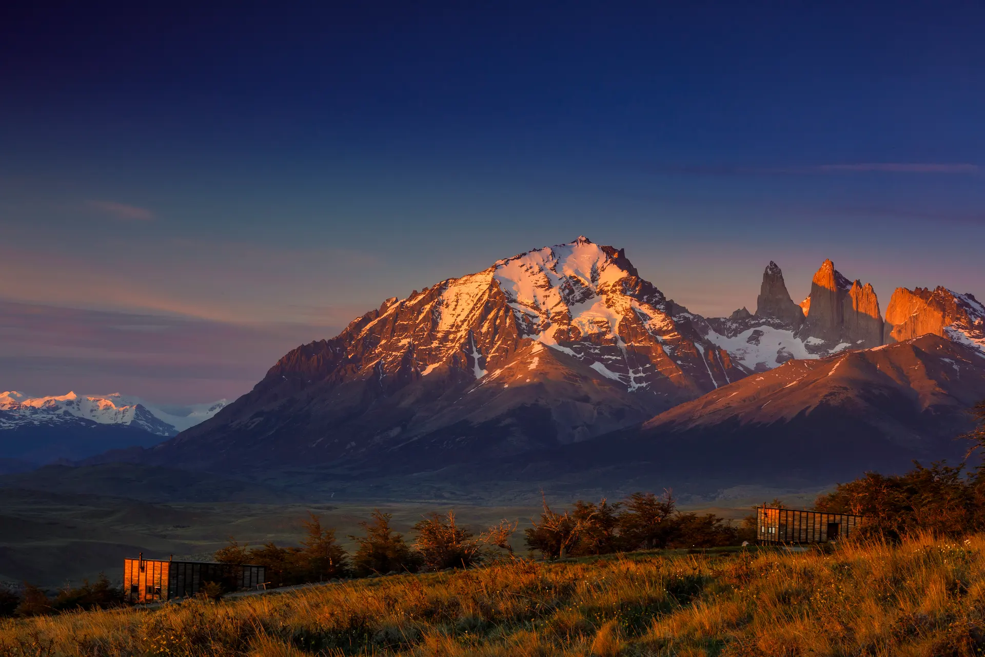 Snow-capped mountain peaks glowing at sunrise, with golden grassland and small cabins in the foreground.