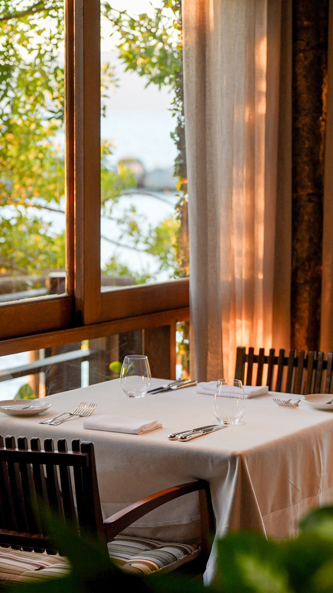 Intimate restaurant table with white linens, glassware, and window-side seating overlooking lush greenery.