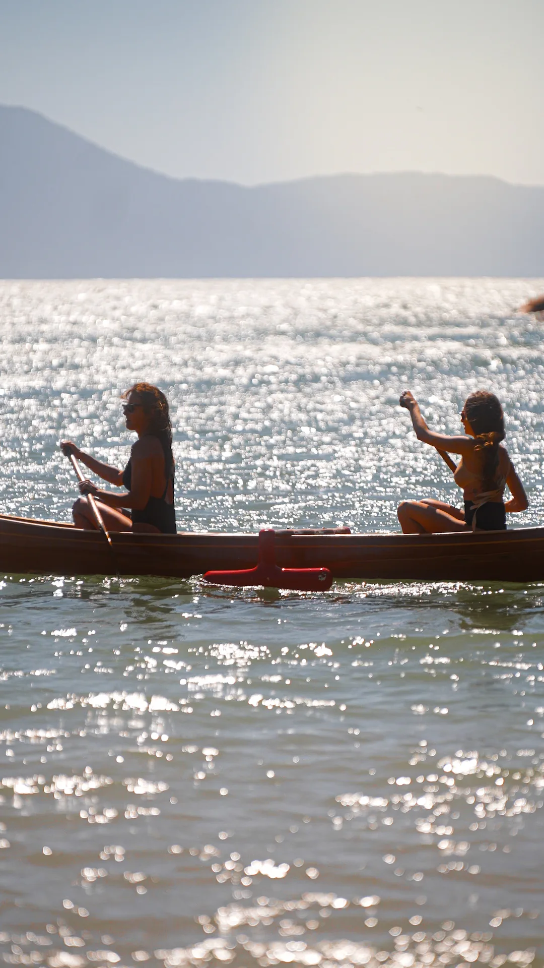 Two people paddling a canoe across shimmering water with mountains in the hazy background.