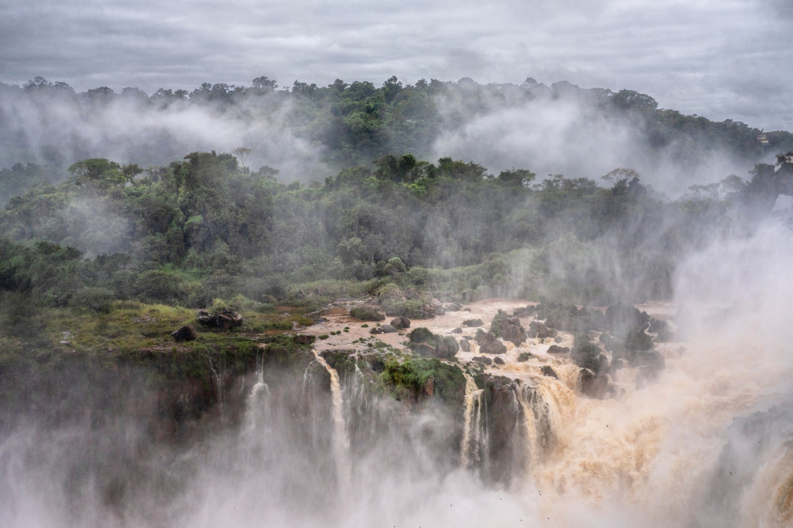 Powerful waterfalls plunging through misty rainforest cliffs, surrounded by dense green foliage under a cloudy sky.