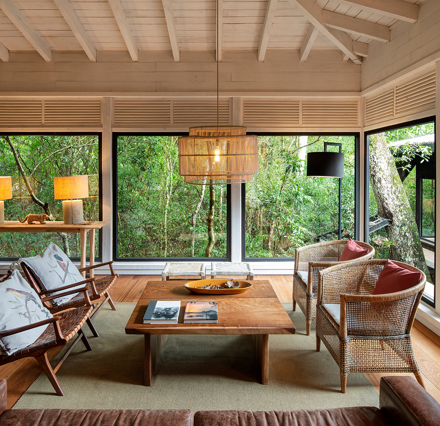Cozy living room with wooden furniture, wicker chairs, and large windows overlooking lush forest greenery.