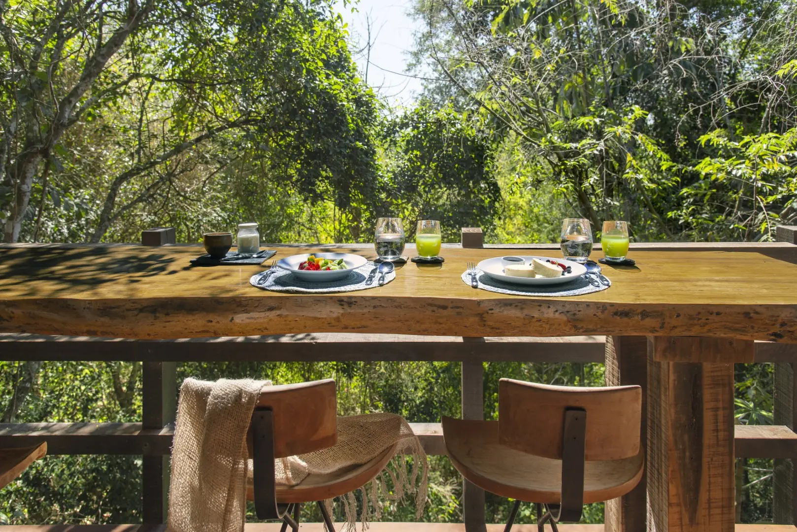 Rustic Outdoor Dining Table Set For Two On A Wooden Deck, Surrounded By Lush Green Forest Foliage.