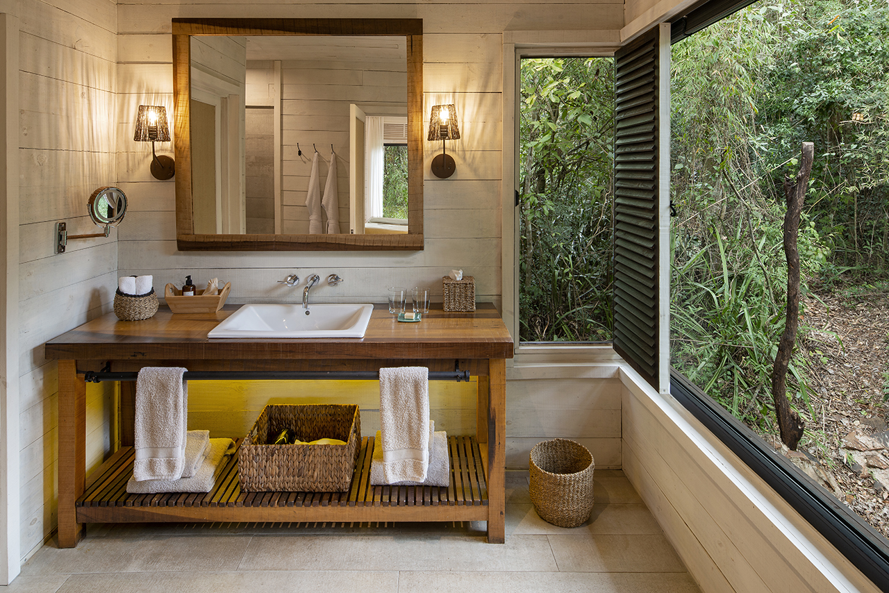 Rustic bathroom with wooden vanity, white sink, towels, and large open window overlooking lush greenery.