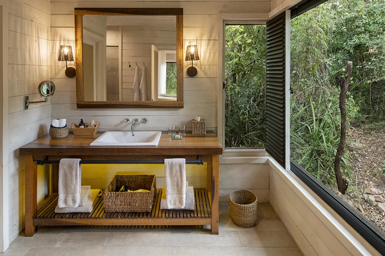 Rustic Bathroom With Wooden Vanity, White Sink, Towels, And Large Open Window Overlooking Lush Greenery.