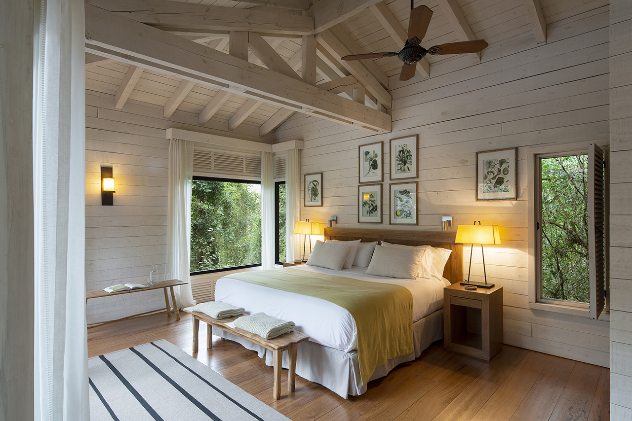 Sunlit rustic bedroom with a king bed, wood beams, botanical wall art, bedside lamps, and forest views through large windows.