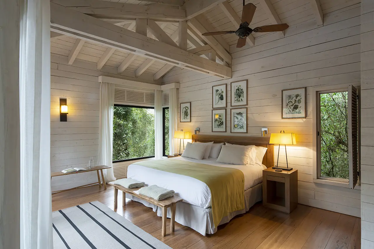 Sunlit Rustic Bedroom With A King Bed, Wood Beams, Botanical Wall Art, Bedside Lamps, And Forest Views Through Large Windows.