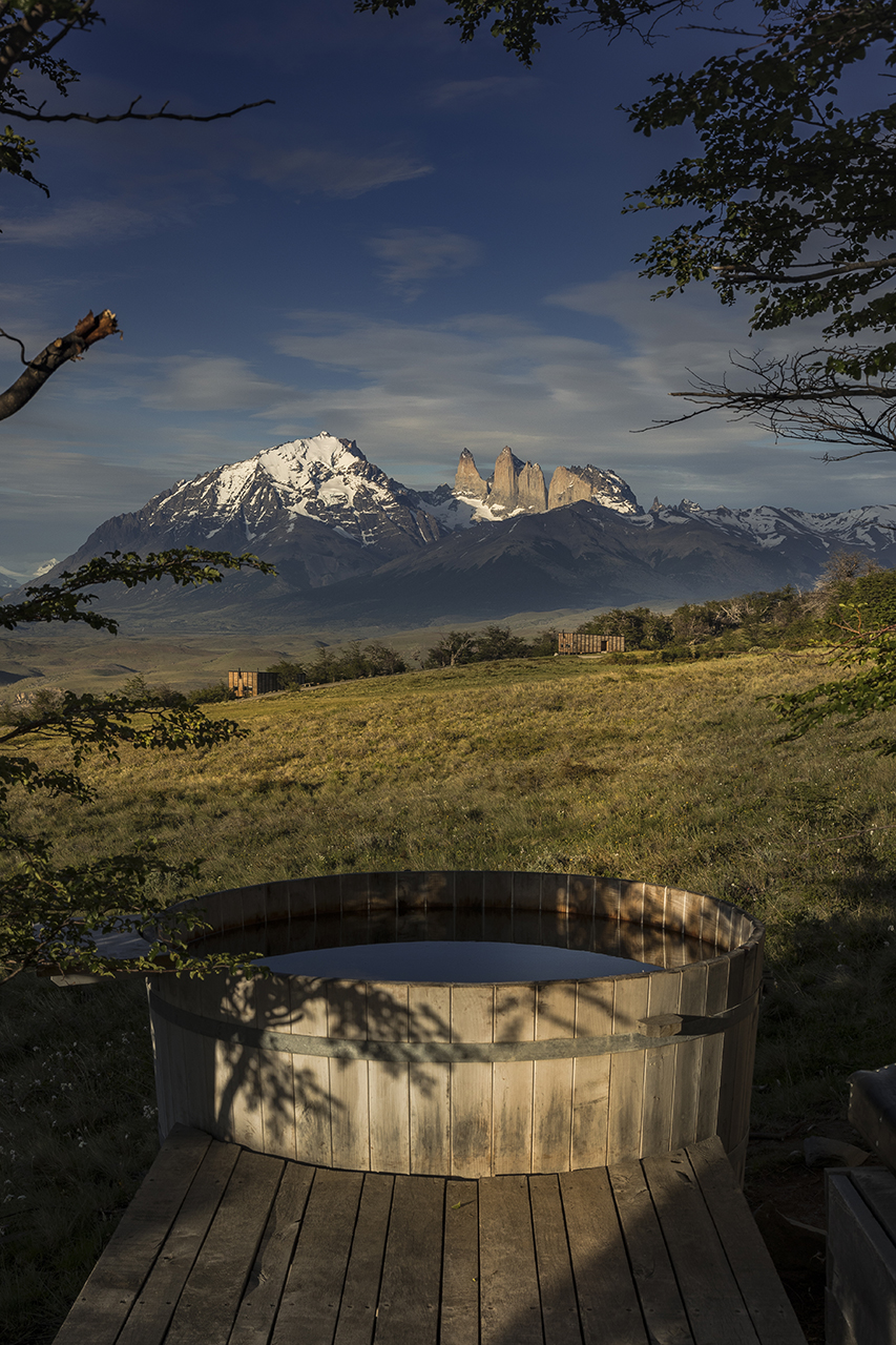 Wooden outdoor soaking tub on a grassy hillside, overlooking dramatic snow-capped mountains beneath a moody blue sky.