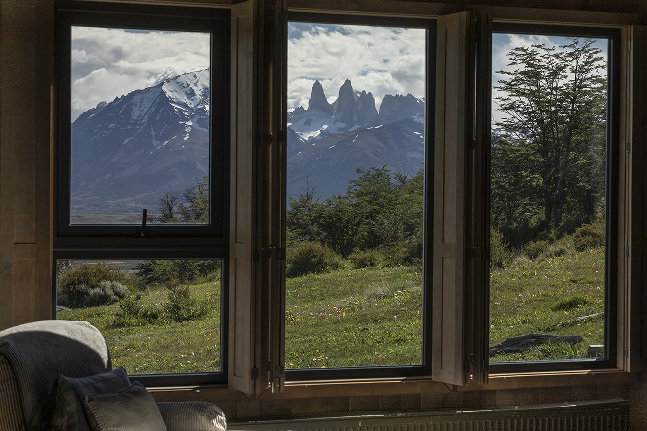 Mountain peaks and green meadow seen through open cabin windows, with trees in the foreground and cloudy sky above.