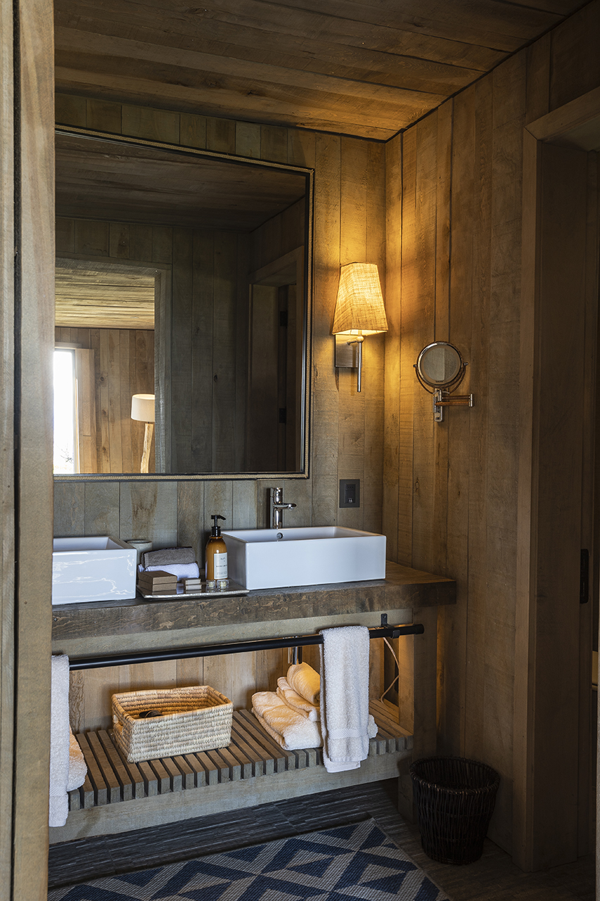 Rustic bathroom with wooden walls, white sink, large mirror, warm lighting, and neatly folded towels.