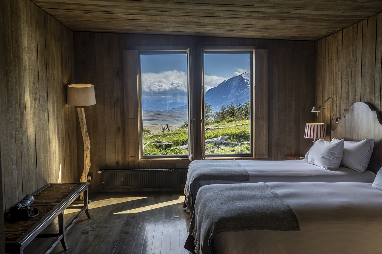 Rustic bedroom with twin beds, wooden walls, and large window overlooking mountains and open landscape.
