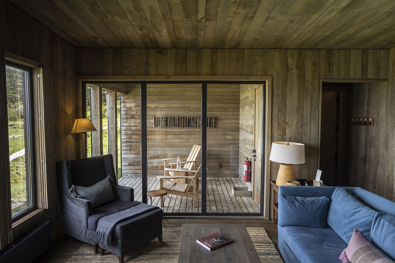 Cozy wood-paneled living room with armchair, sofa, sliding glass doors, and covered deck with chairs outside.