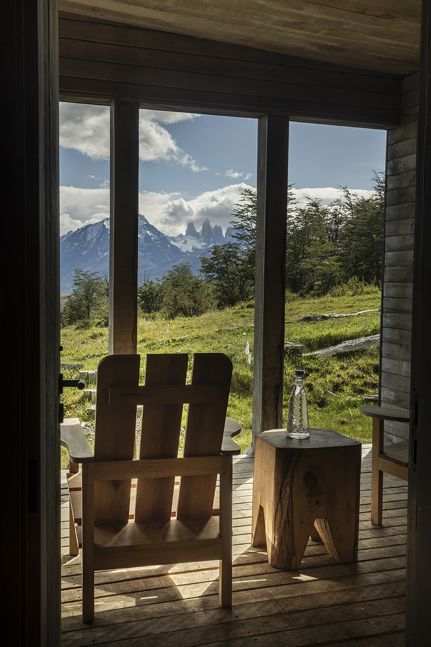 Wooden chairs on a cabin porch overlooking grassy hills and dramatic snowcapped mountains through an open doorway.