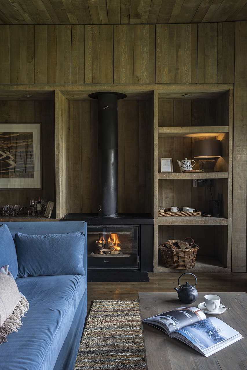 Cozy cabin interior with wood-paneled walls, lit fireplace, blue sofa, coffee table with book and tea set.