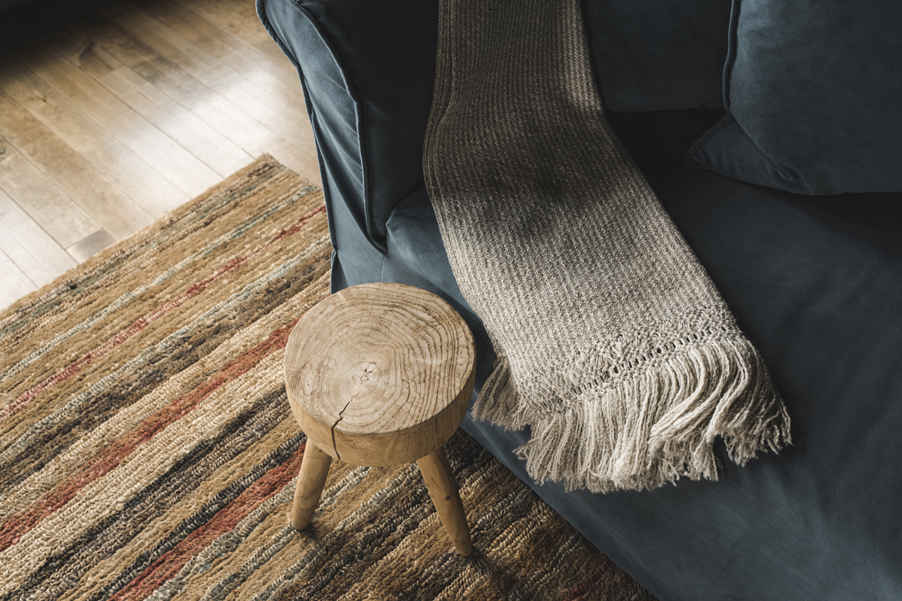 Cozy living room corner with a blue sofa, textured throw blanket, rustic wooden stool, and woven rug on wood floors.