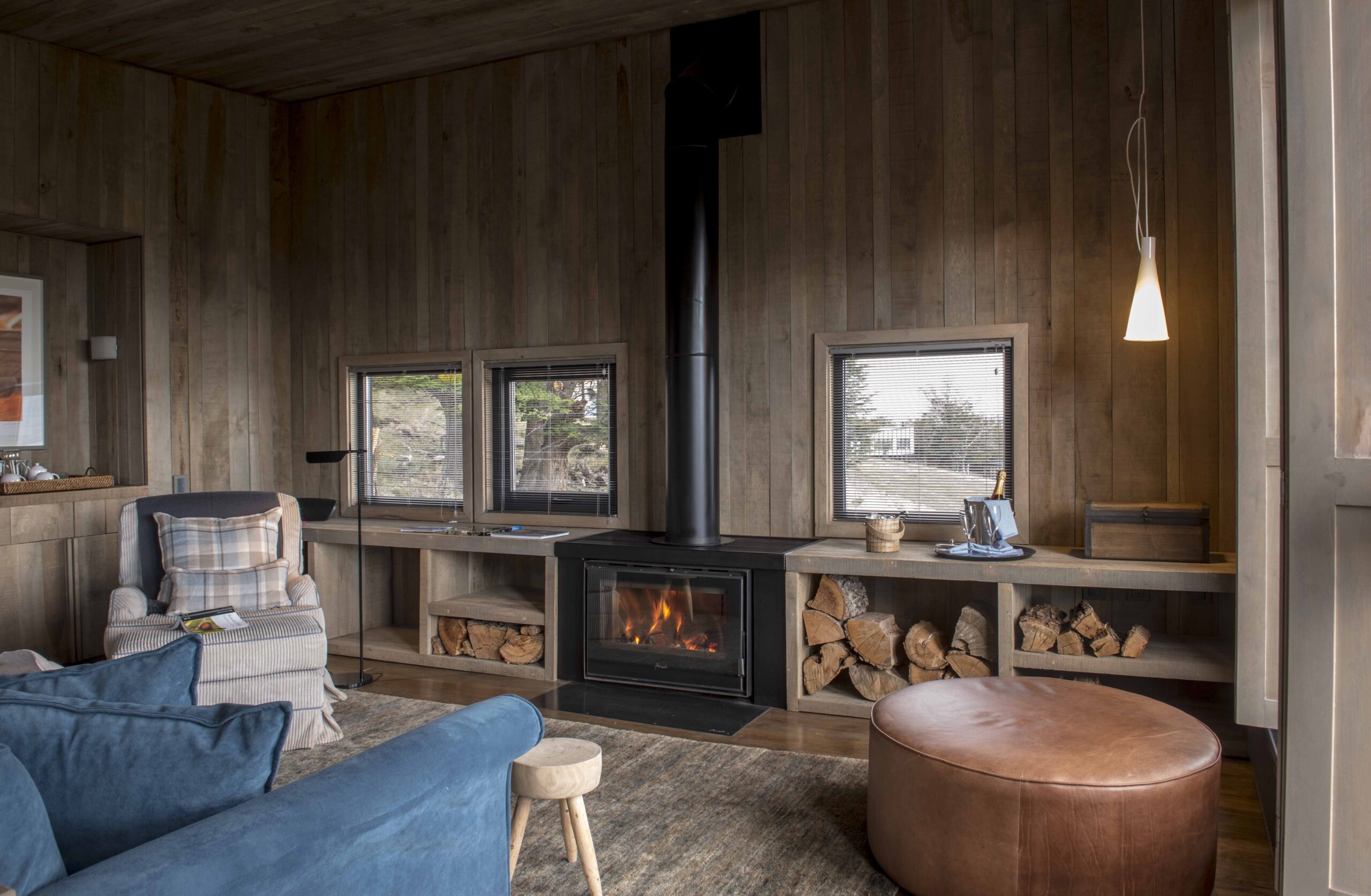 Warm wood-paneled living room with fireplace, stacked firewood, cozy seating, soft lighting, and small windows.