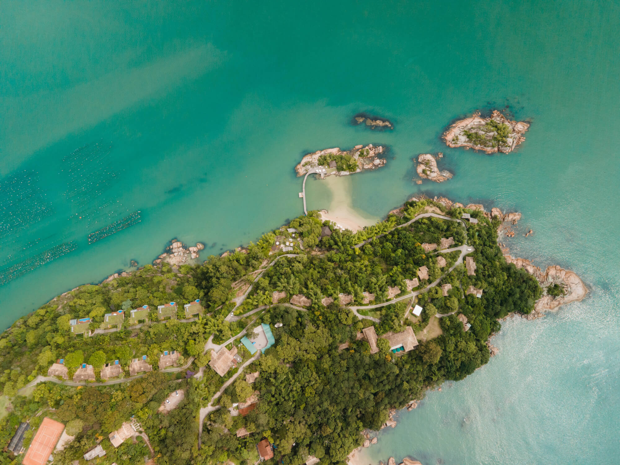 Aerial view of a lush island resort with villas, winding paths, turquoise water, and small rocky islets nearby.