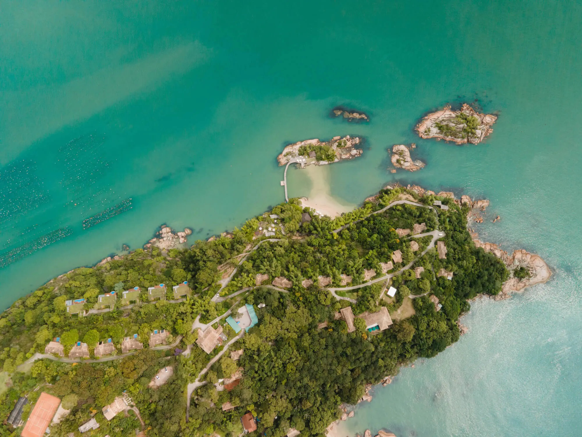 Aerial View Of A Lush Island Resort With Villas, Winding Paths, Turquoise Water, And Small Rocky Islets Nearby.