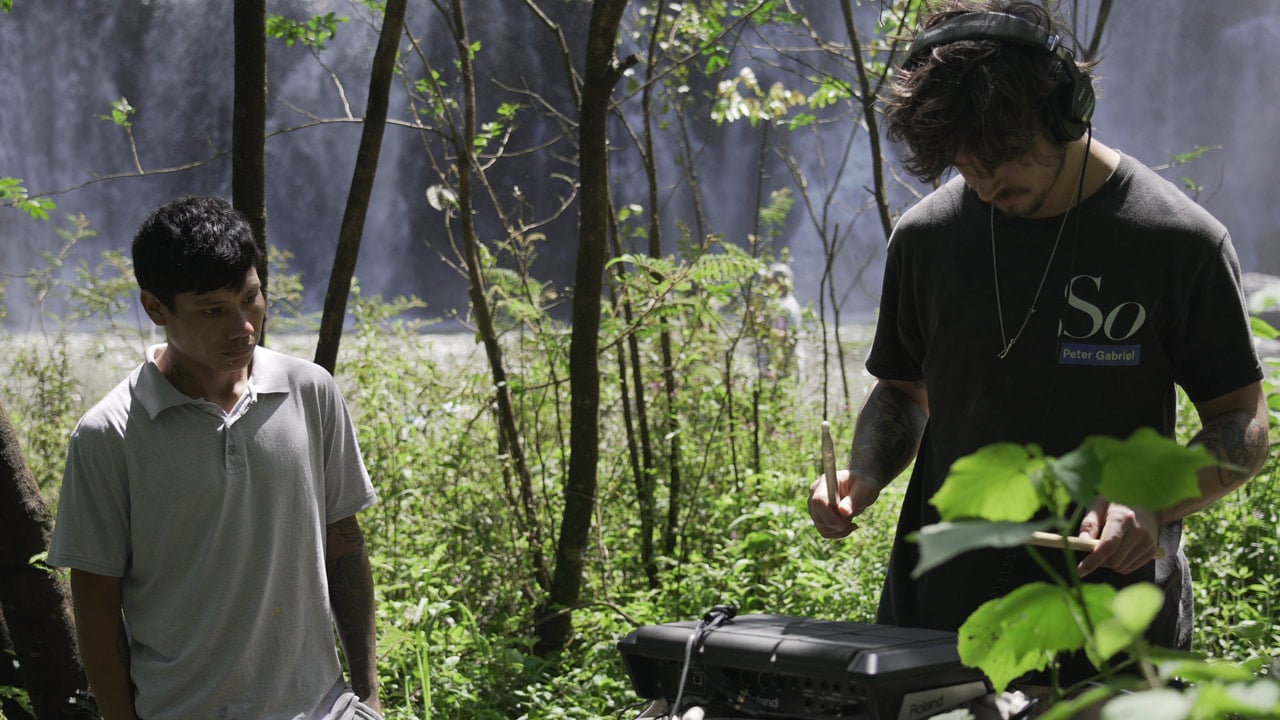 Man playing an electronic drum pad in a forest while another watches.