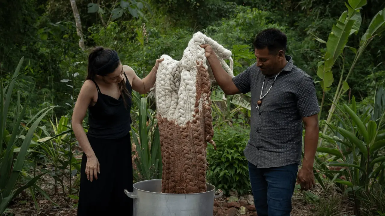 Two people holding a large piece of dyed fabric over a pot outdoors, surrounded by lush vegetation.