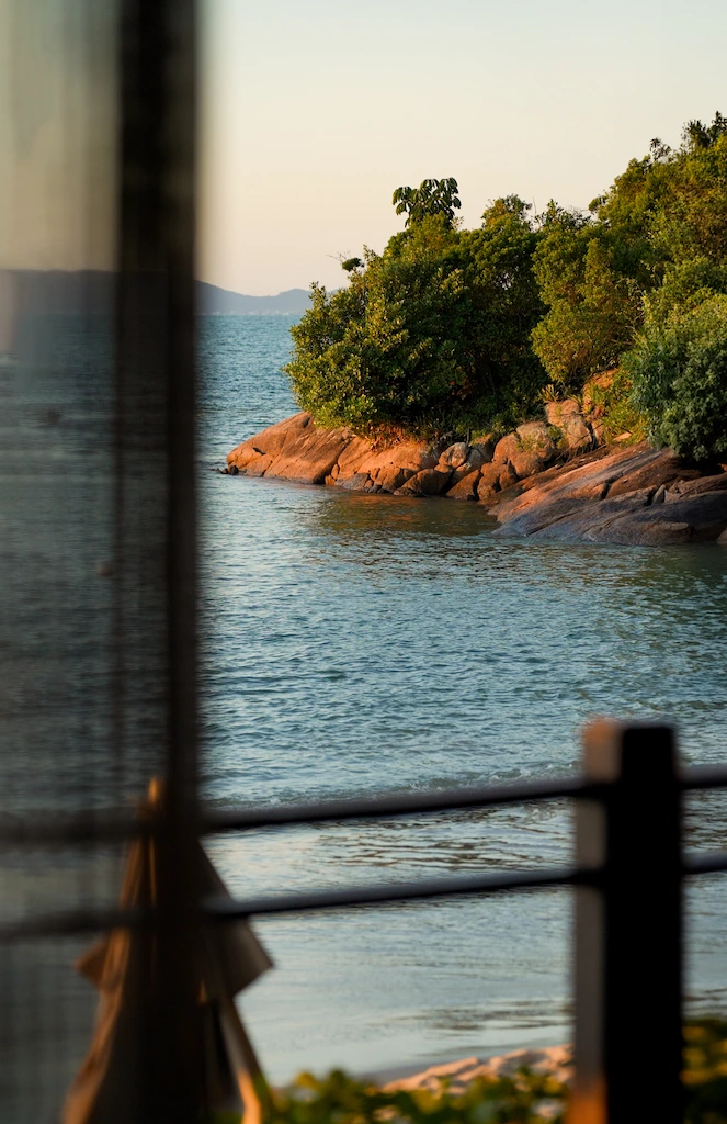 Rocky shoreline with lush greenery beside calm sea, viewed from a shaded terrace at sunset.