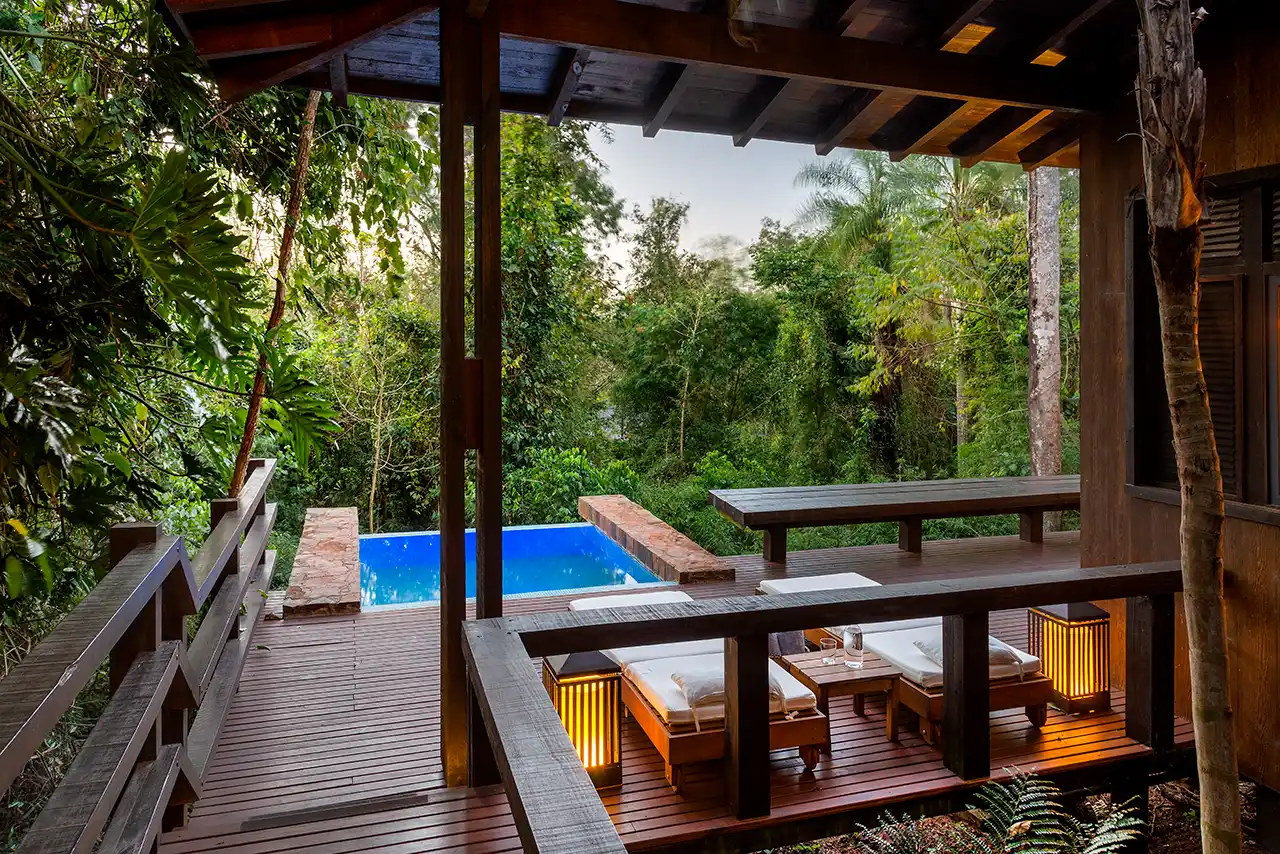 Wooden deck with lounge chairs and warm lantern lighting surrounded by dense greenery and a small pool.