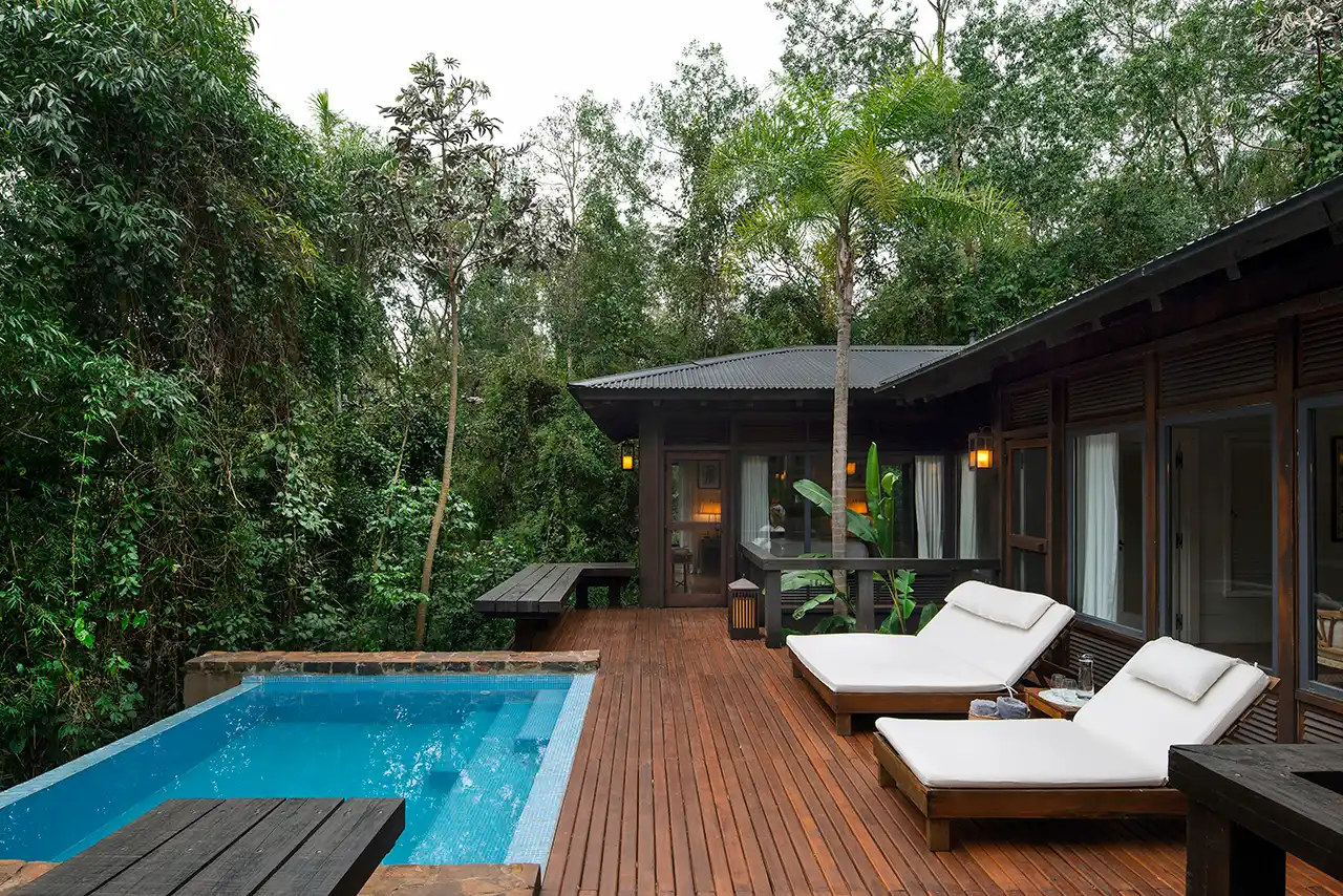 Wooden deck with two white lounge chairs and a pool surrounded by dense forest.