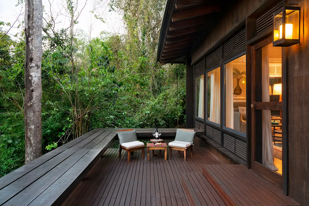 Wooden deck with two cushioned chairs and a small table beside a modern house surrounded by greenery.