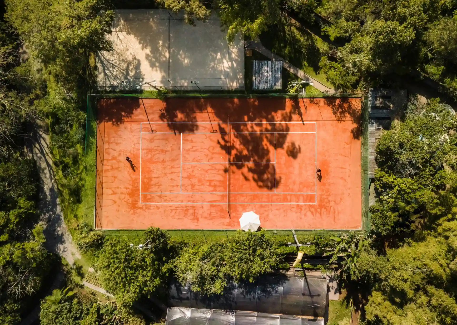 Aerial view of a clay tennis court surrounded by dense green trees with two players on court.