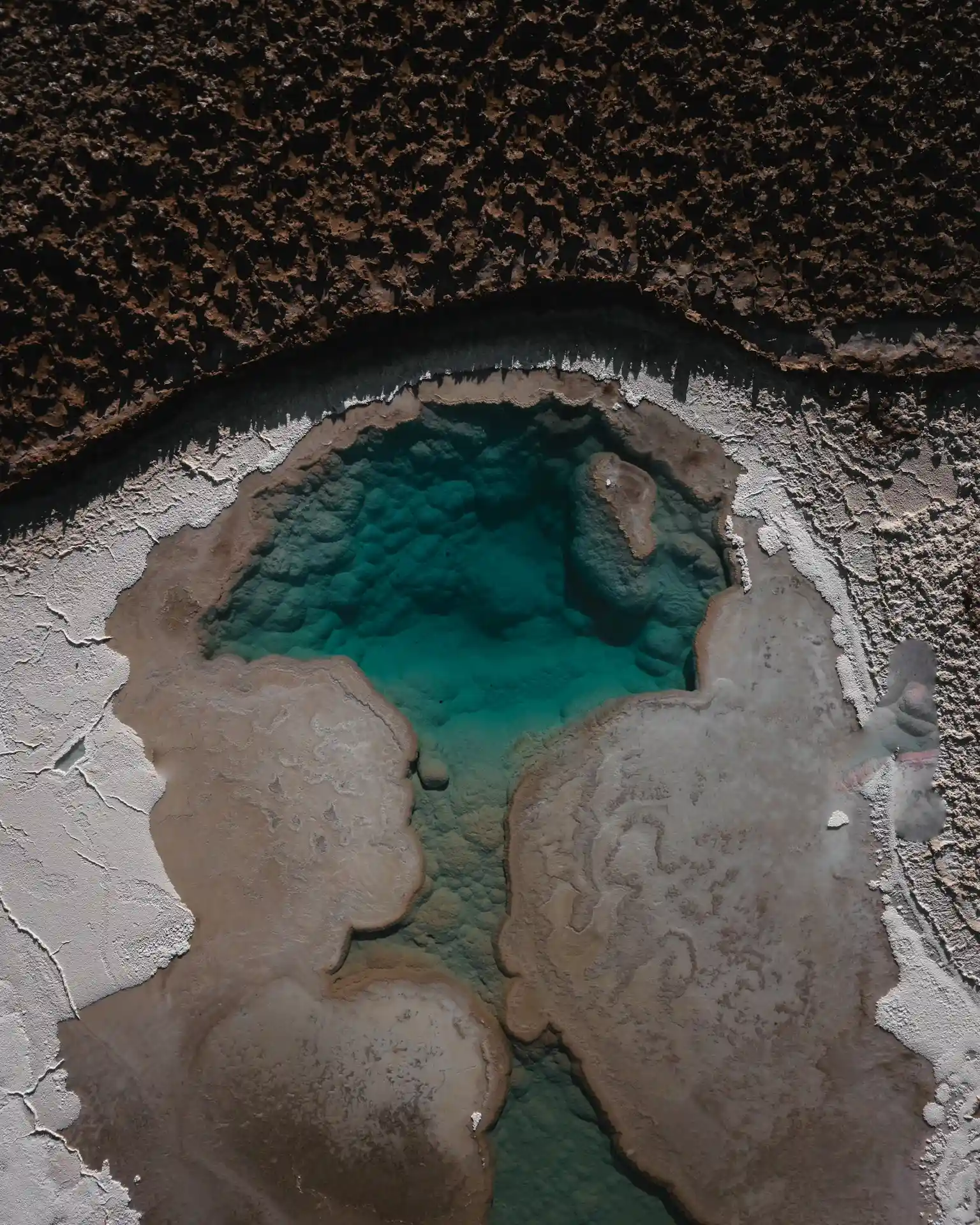 Aerial view of a turquoise mineral pool surrounded by textured salt flats and dark rocky terrain.
