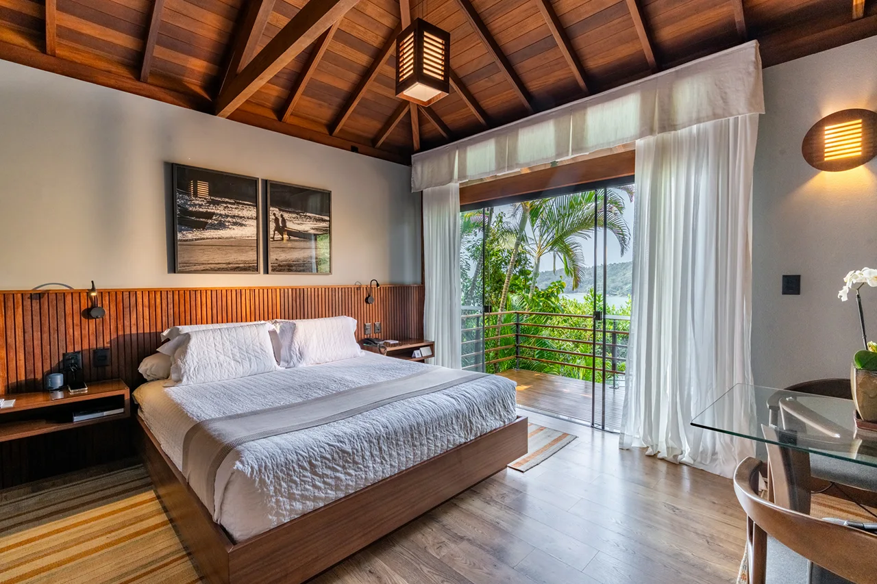 Tropical bedroom with a large bed, vaulted ceiling, and glass doors opening to a balcony with palm‑tree views.