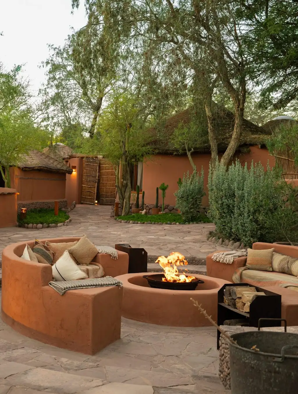 Outdoor fire pit surrounded by curved adobe benches with cushions, set among rustic thatched buildings and greenery.