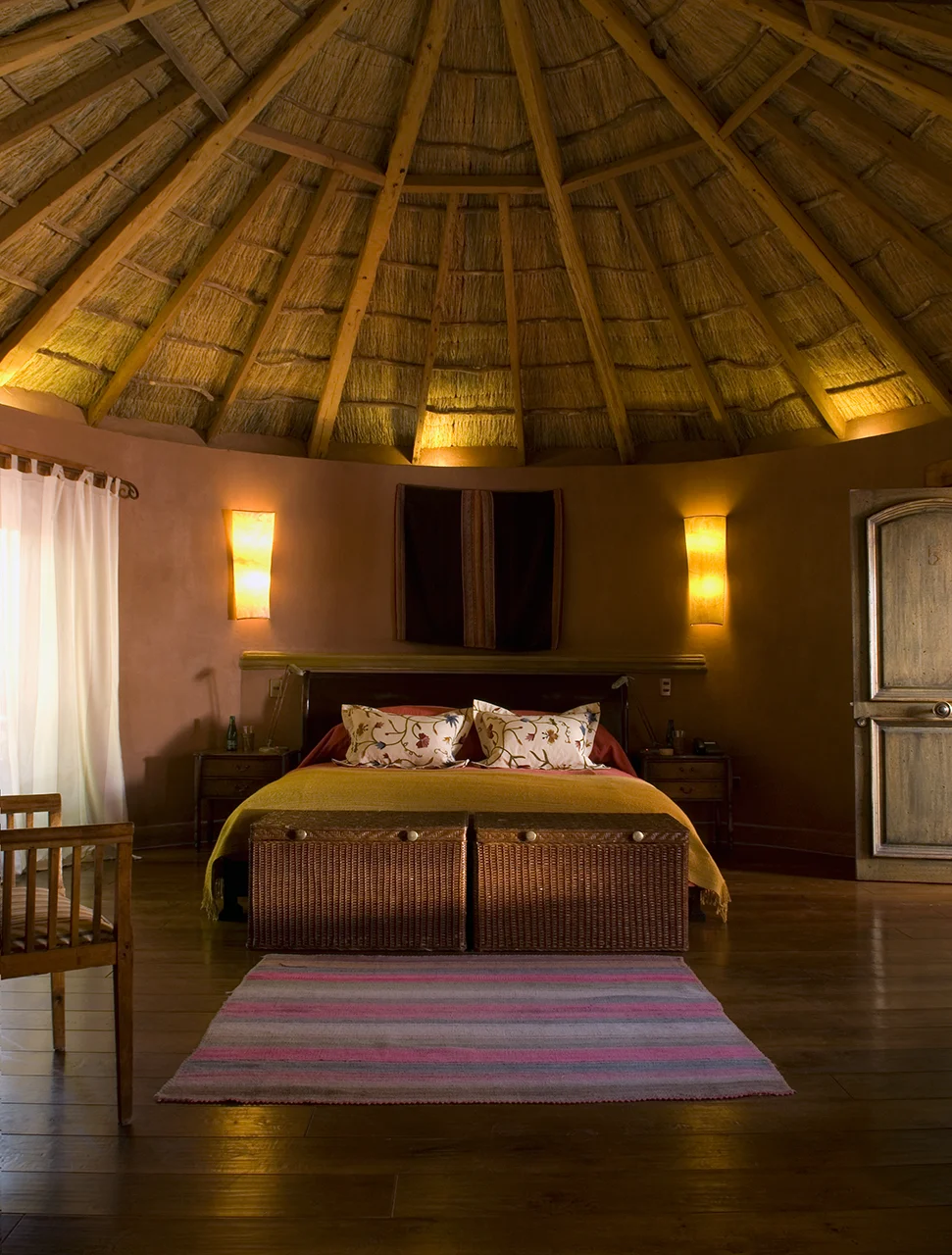Rustic bedroom with a thatched roof, double bed with floral pillows, woven baskets, and warm wooden accents.
