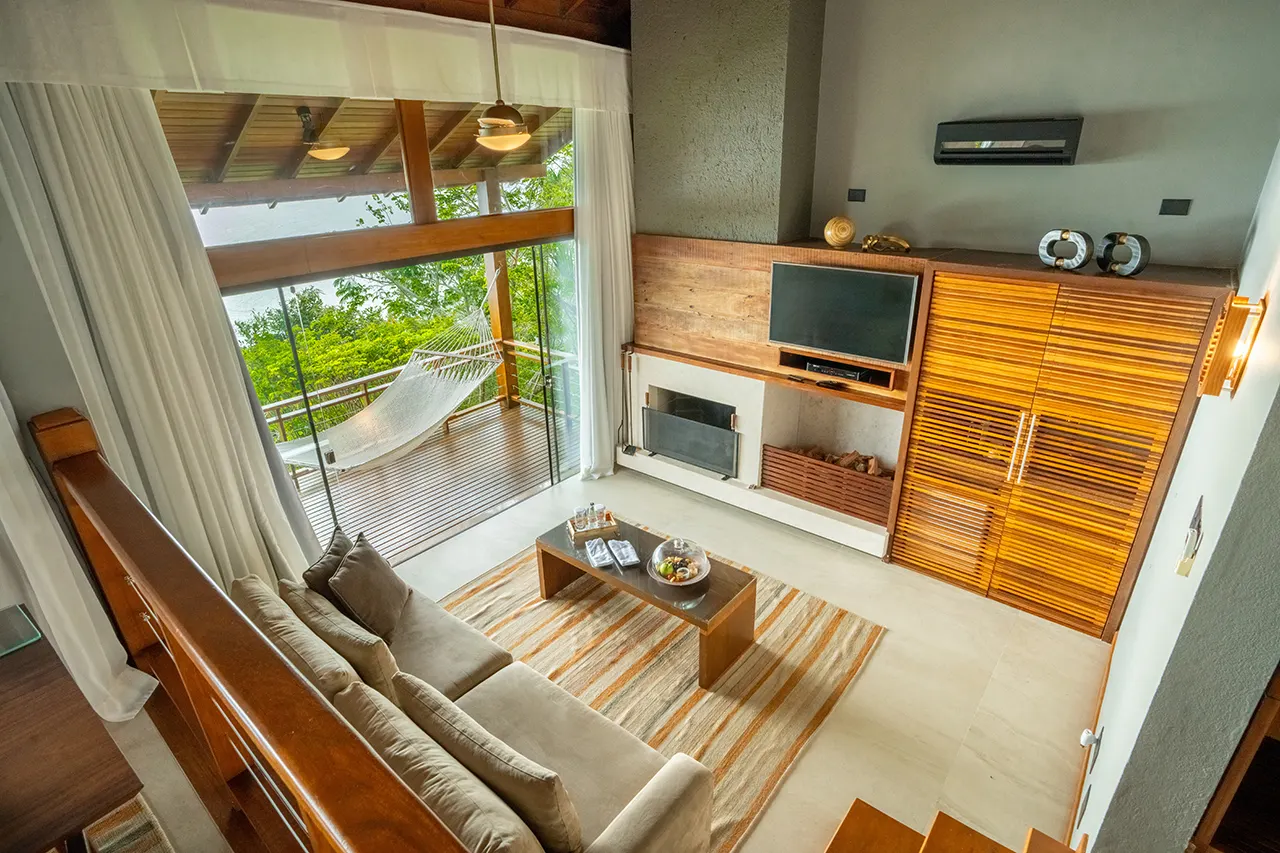 Living room with a beige sectional sofa, striped rug, fireplace and TV, opening to a deck with a hammock and greenery.