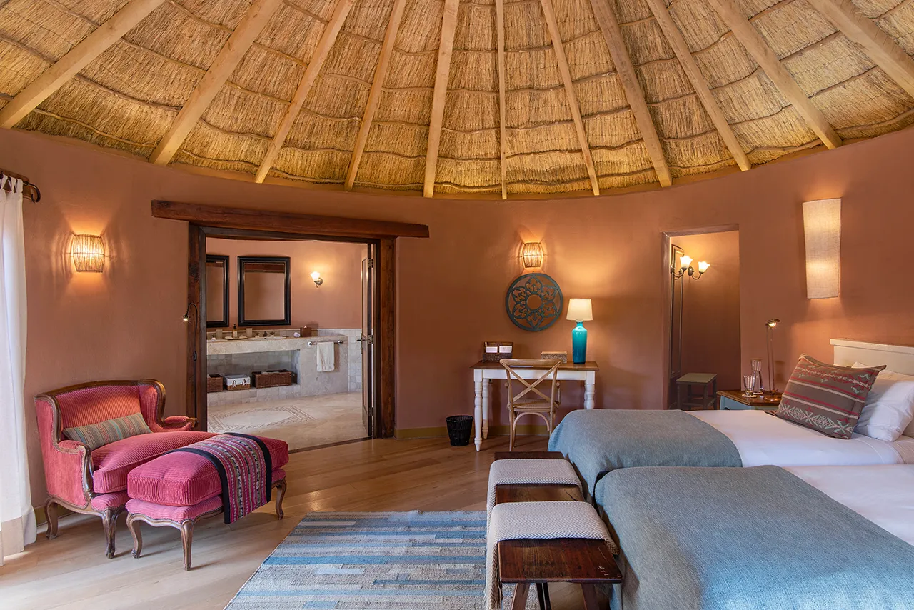Cozy bedroom with two blue‑topped beds, thatched ceiling, red armchair, and a doorway leading to a double‑sink bathroom.
