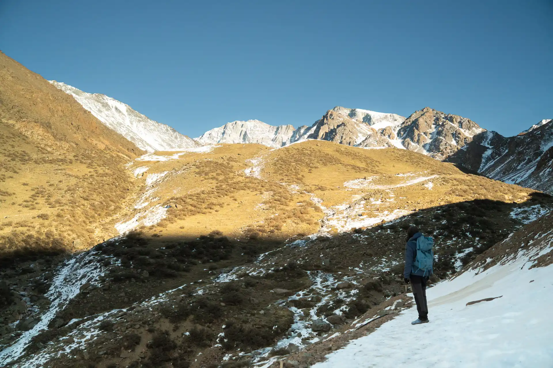 Hiker in a blue jacket standing on a snowy trail with grassy slopes and rugged, snow‑capped mountains under a clear sky.