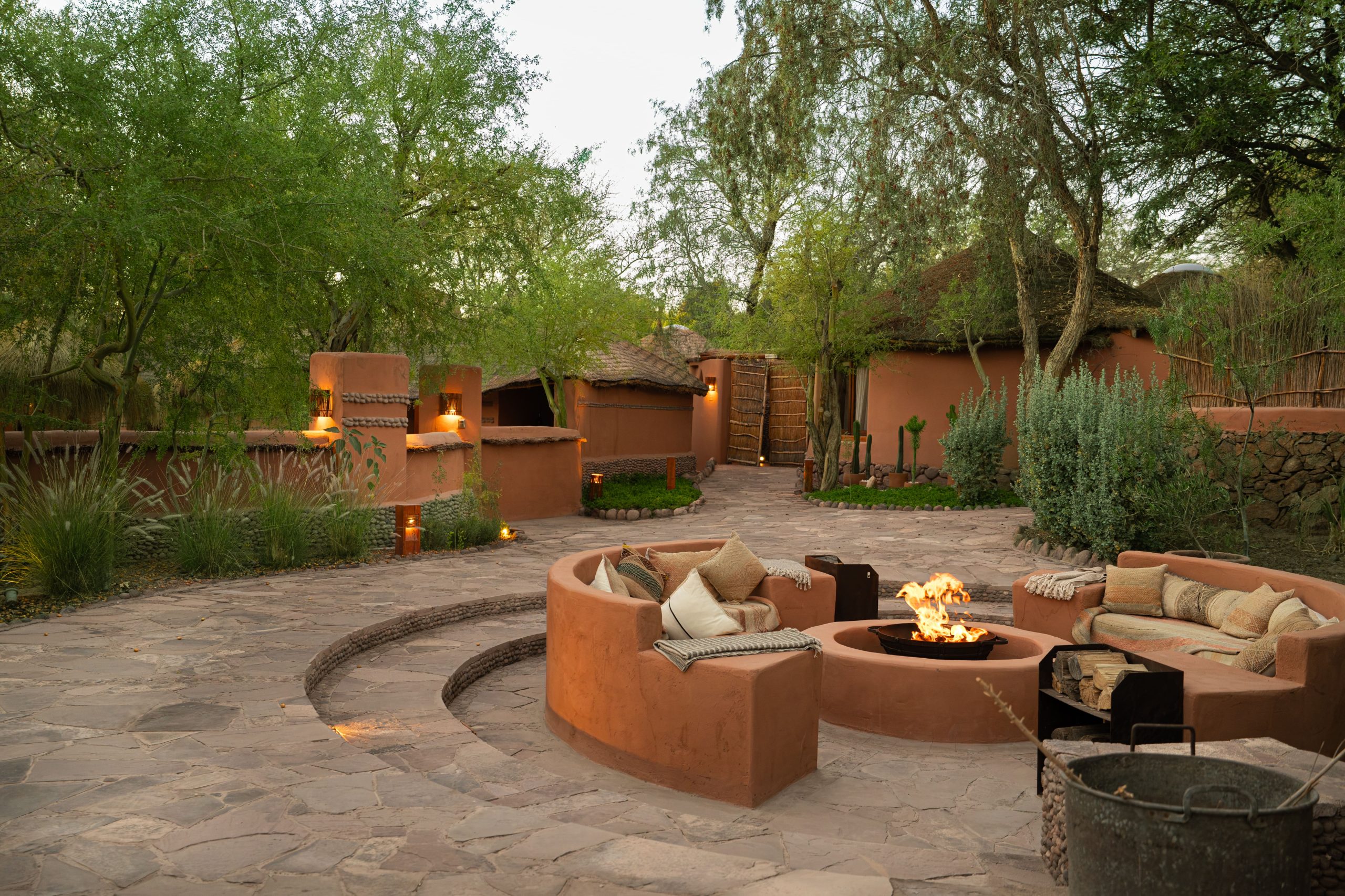 Circular adobe patio with cushioned seating around a glowing fire pit, framed by greenery and warm lights.