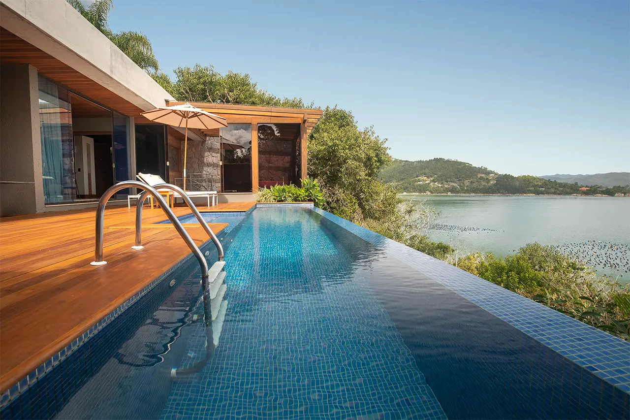 Infinity pool and wooden deck overlooking calm water and green hills beside a modern glass‑fronted villa.