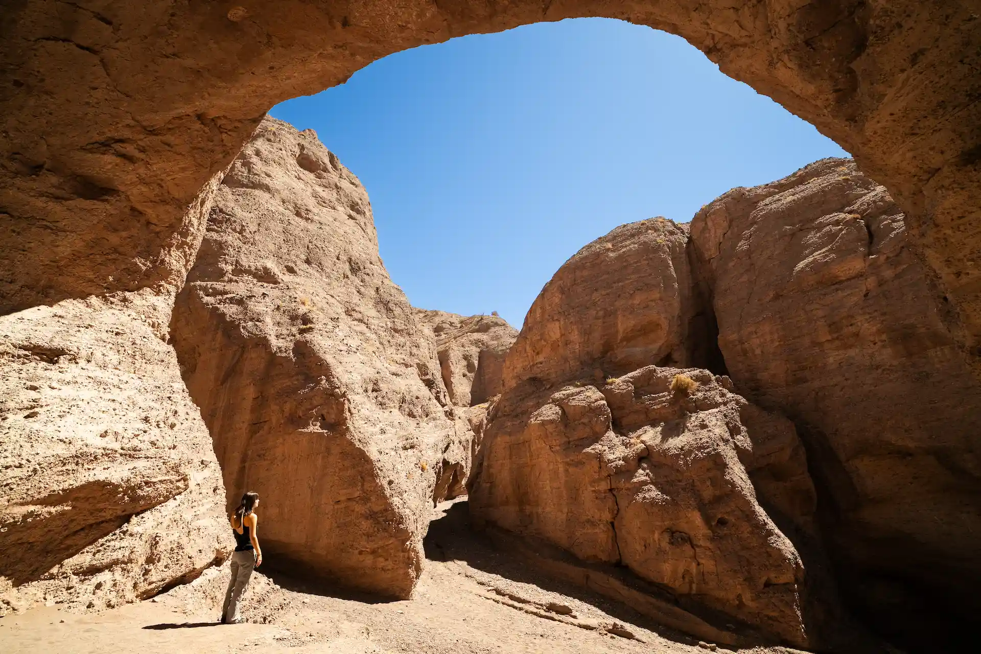 Person standing beneath a towering desert rock arch against a clear blue sky.