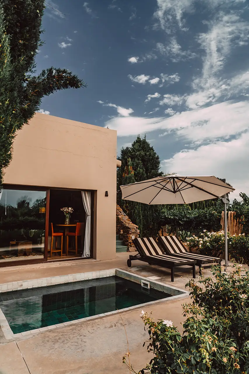 Small pool with striped loungers and umbrella beside a modern suite framed by lush greenery.