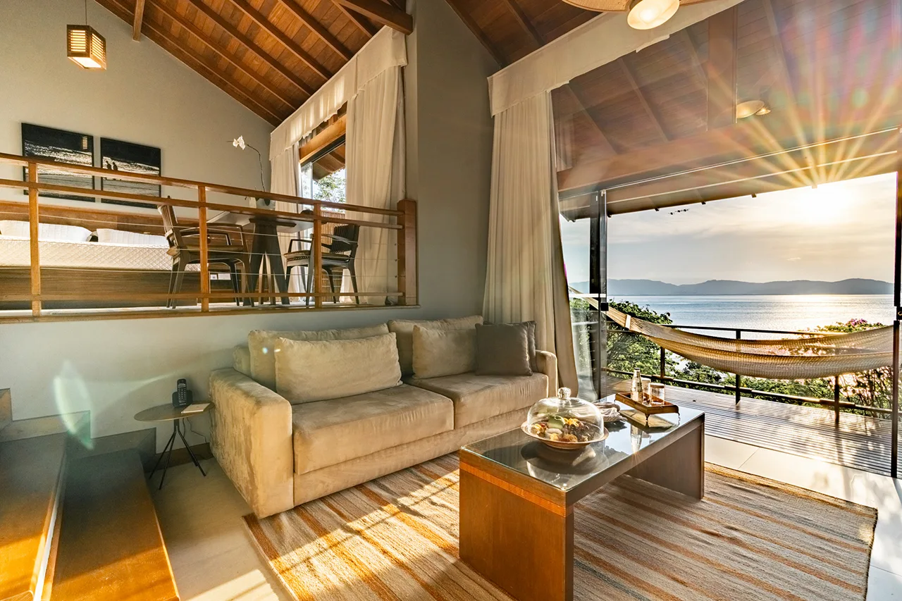 Split‑level suite with sofa, hammock balcony, and sunset views over calm water and distant mountains.