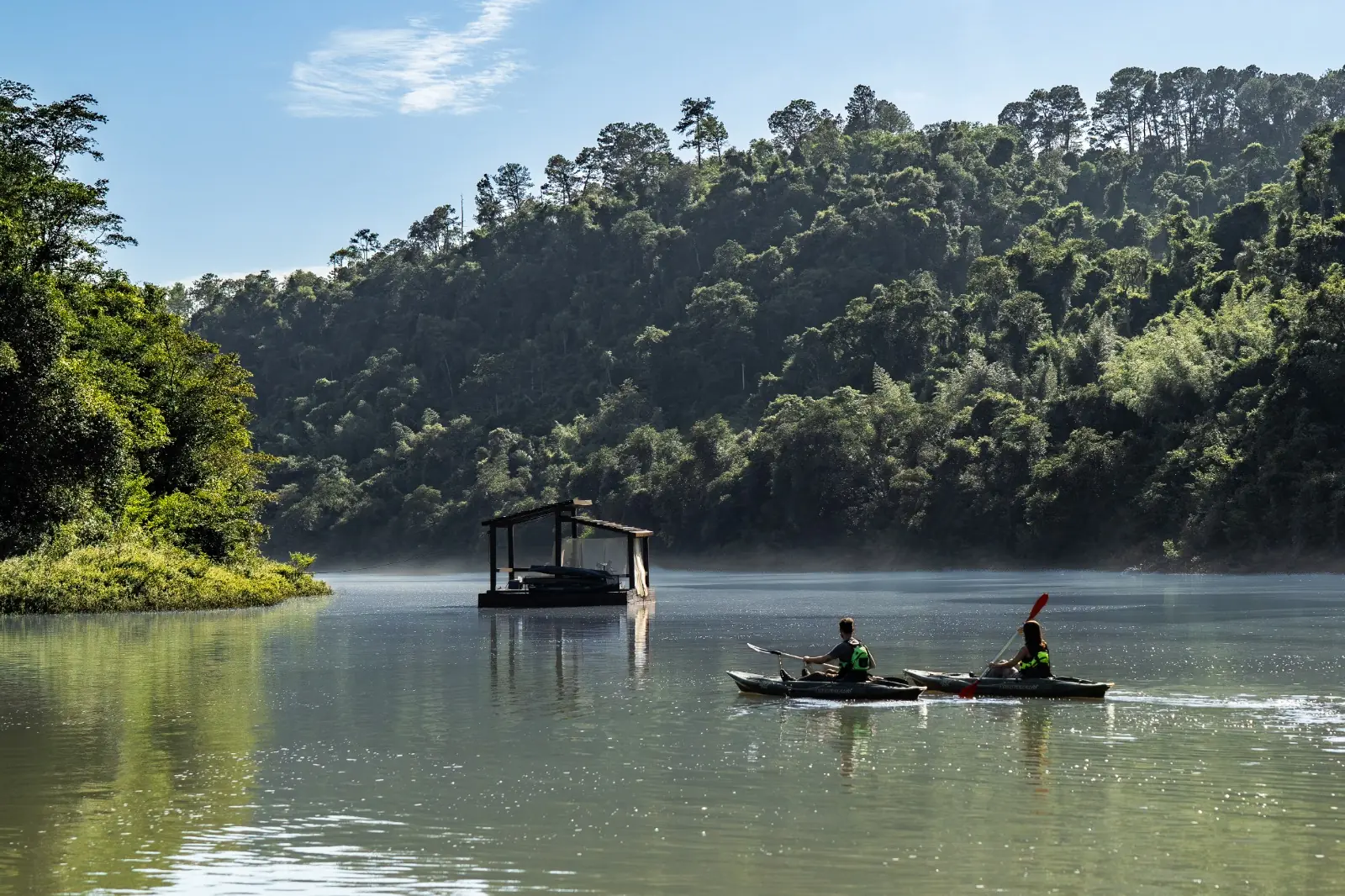 Two people kayaking on a calm river surrounded by lush forest and morning light