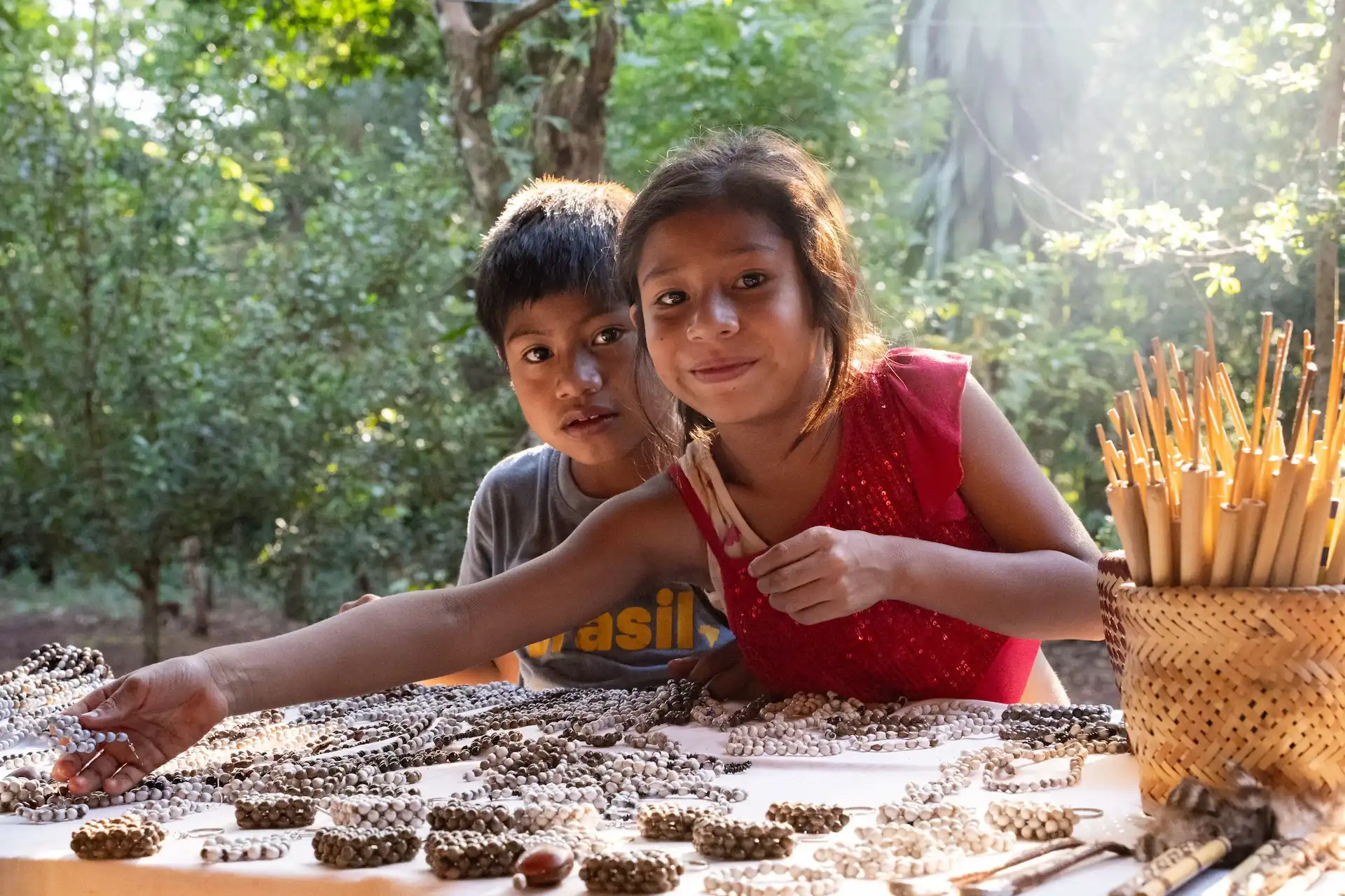 Two children at an outdoor market table, reaching for handmade bead jewelry displayed in sunlight among forest surroundings.
