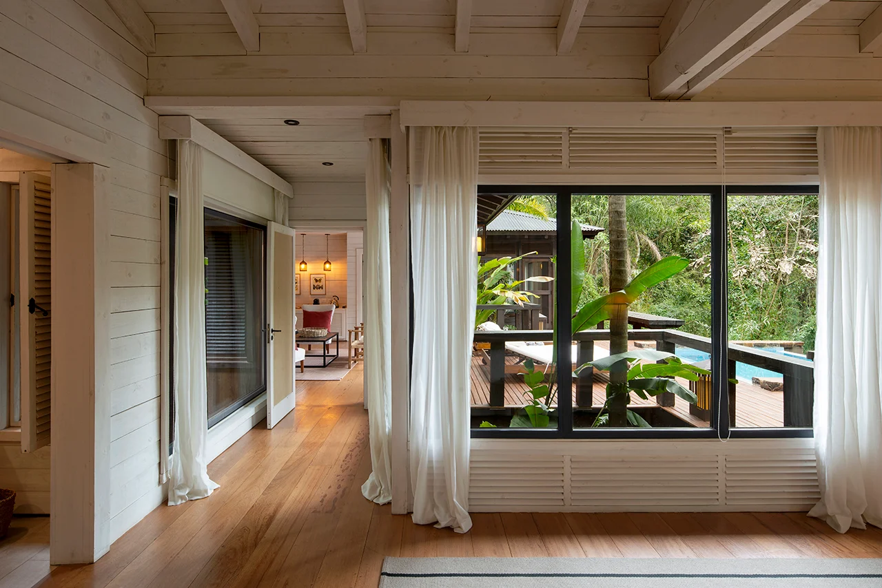 Bright wooden interior with large windows, soft curtains, and views of a tropical terrace and pool