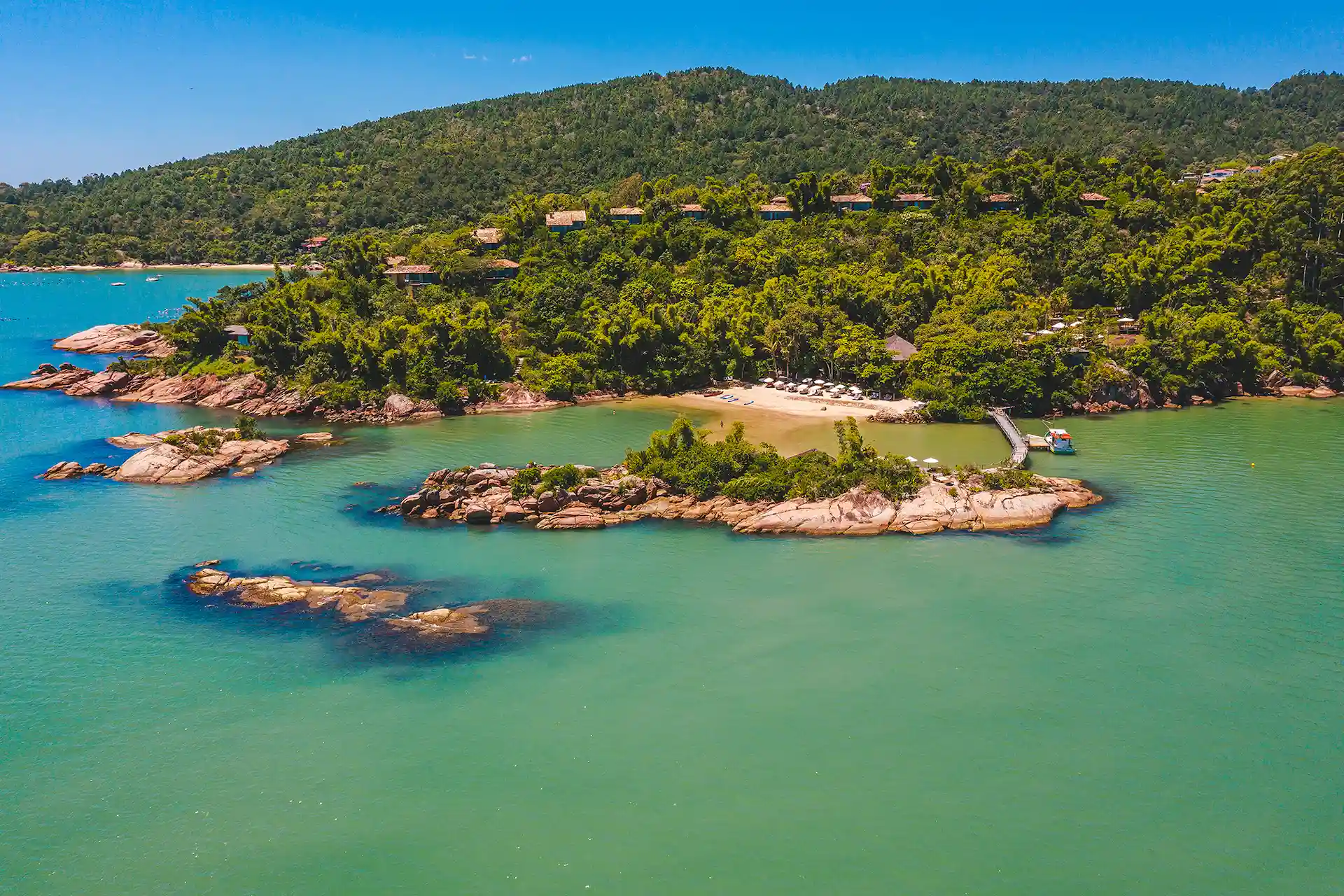 Aerial View Of A Lush Tropical Island With Turquoise Waters, Rocky Shores, Beach Loungers, And Hillside Villas.