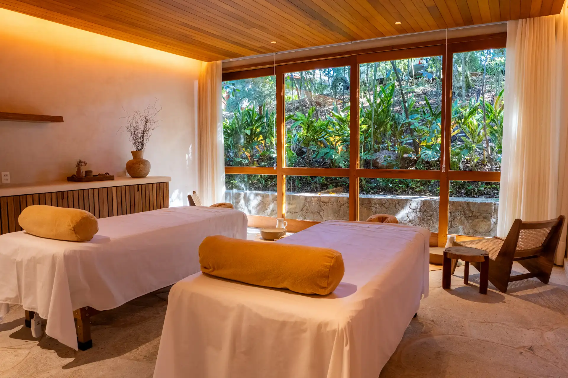 Two massage tables with rolled towels in a tranquil spa room, warm lighting, and large windows facing greenery.