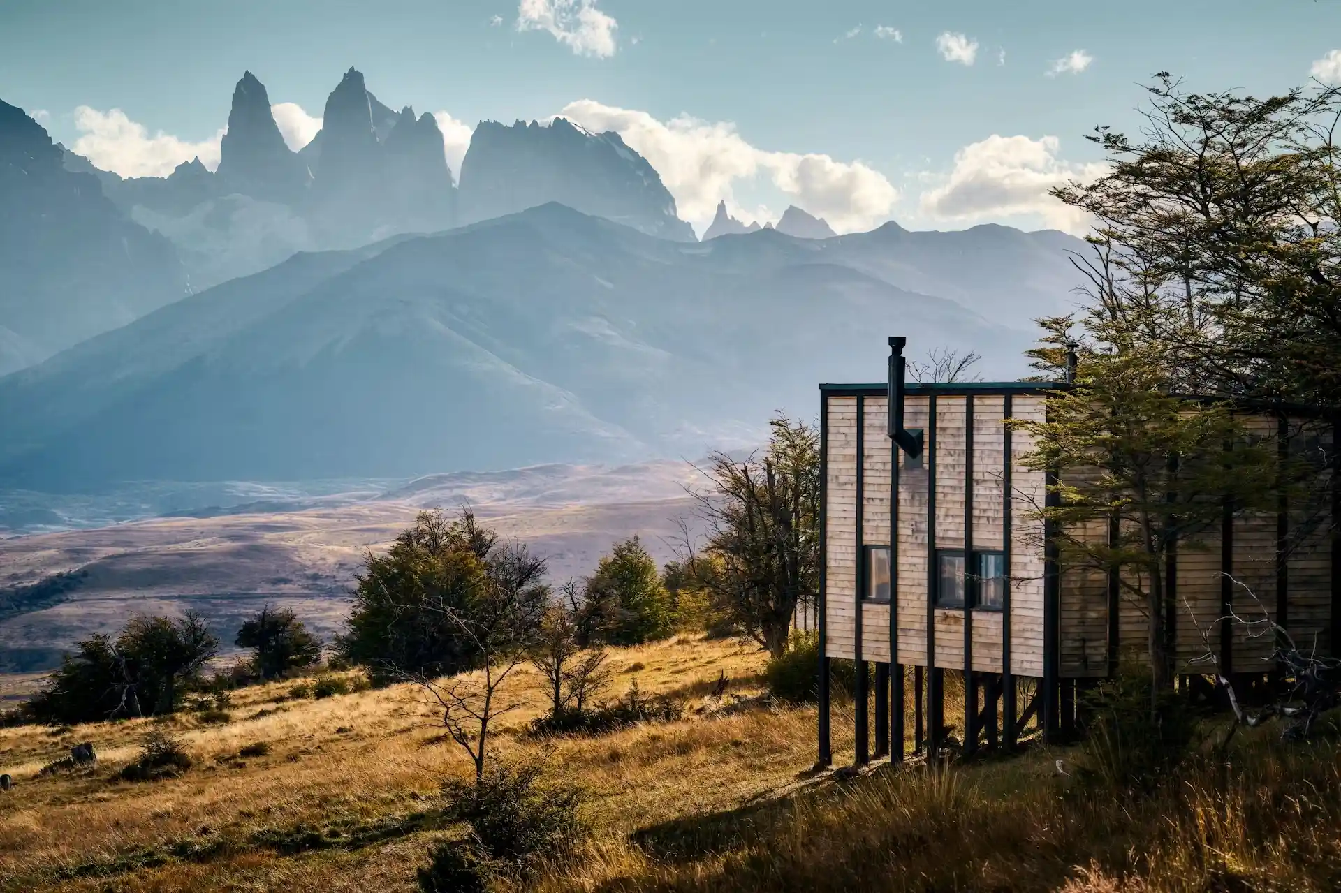 Modern cabin on stilts overlooking golden grasslands with dramatic mountain peaks and a blue sky in the distance.