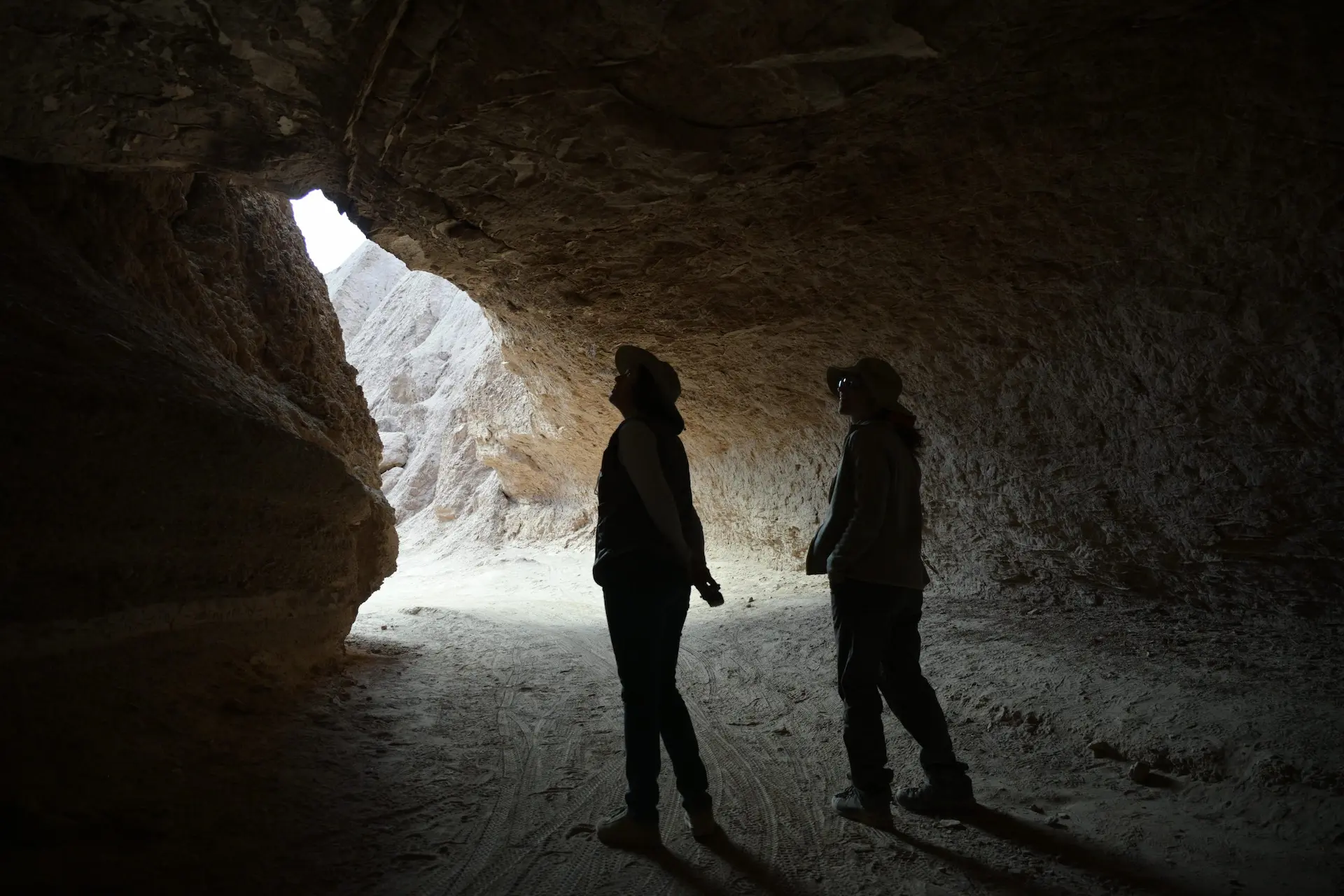 Two people exploring a dark cave, silhouetted against the sunlight streaming in from the entrance, with rugged rock walls surrounding them.