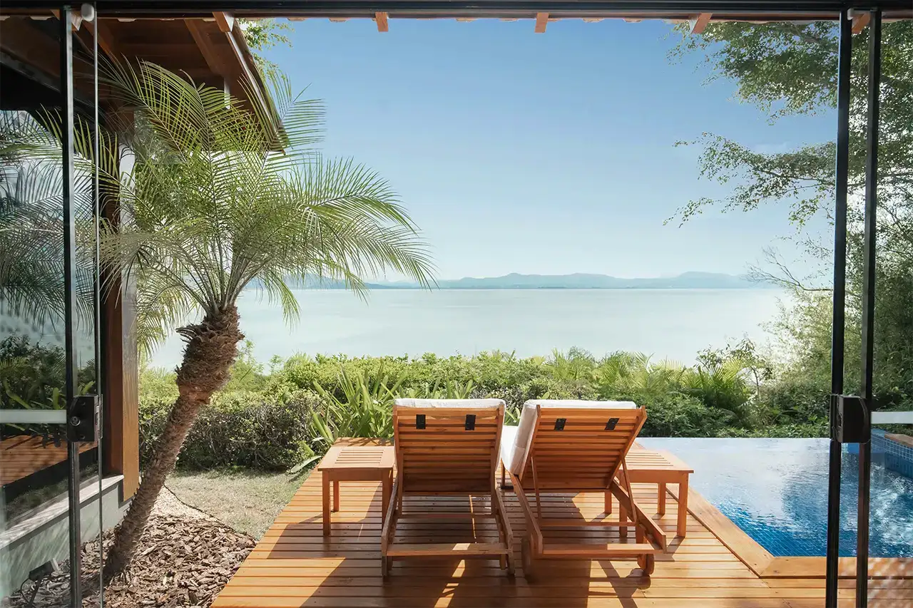 Two sun loungers on a wooden deck by a private pool overlooking the ocean with lush greenery and clear blue sky