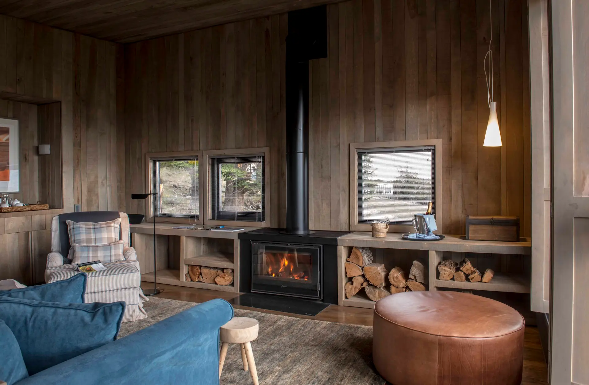 Cozy wooden cabin interior with fireplace seating area large windows and stacked firewood creating a warm relaxing atmosphere